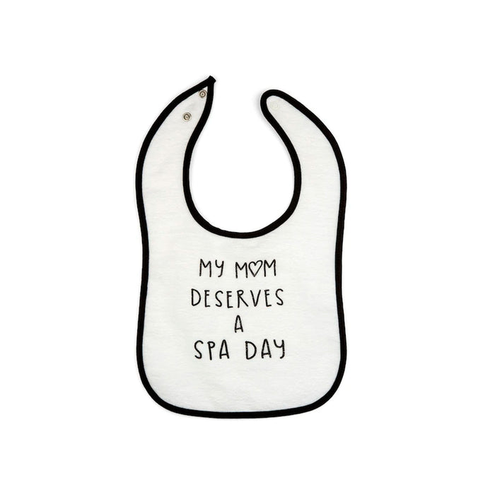 White baby bib with black trim and the phrase "My mom deserves a spa day" printed in simple black lettering, featuring the product name Mom Deserves A Spa Day Baby Bib | Lucky Owl.